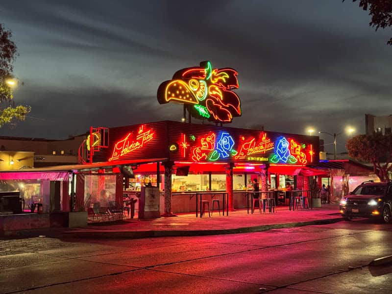 La Fabrica del Taco restaurant lit up with neon lights under a grey sky.